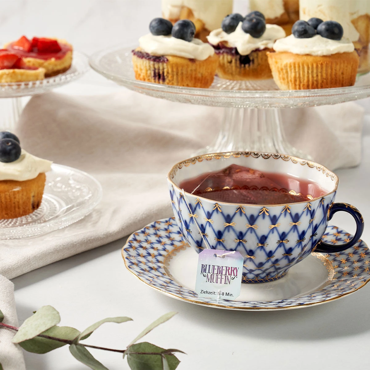 Eine gemusterte Teetasse mit Tee steht auf einer passenden Untertasse neben einem Sweeteas „Blueberry Muffin“-Teebeutel. Im Hintergrund sind Cupcakes mit Zuckerguss und Blaubeeren auf Glasständern ausgestellt.