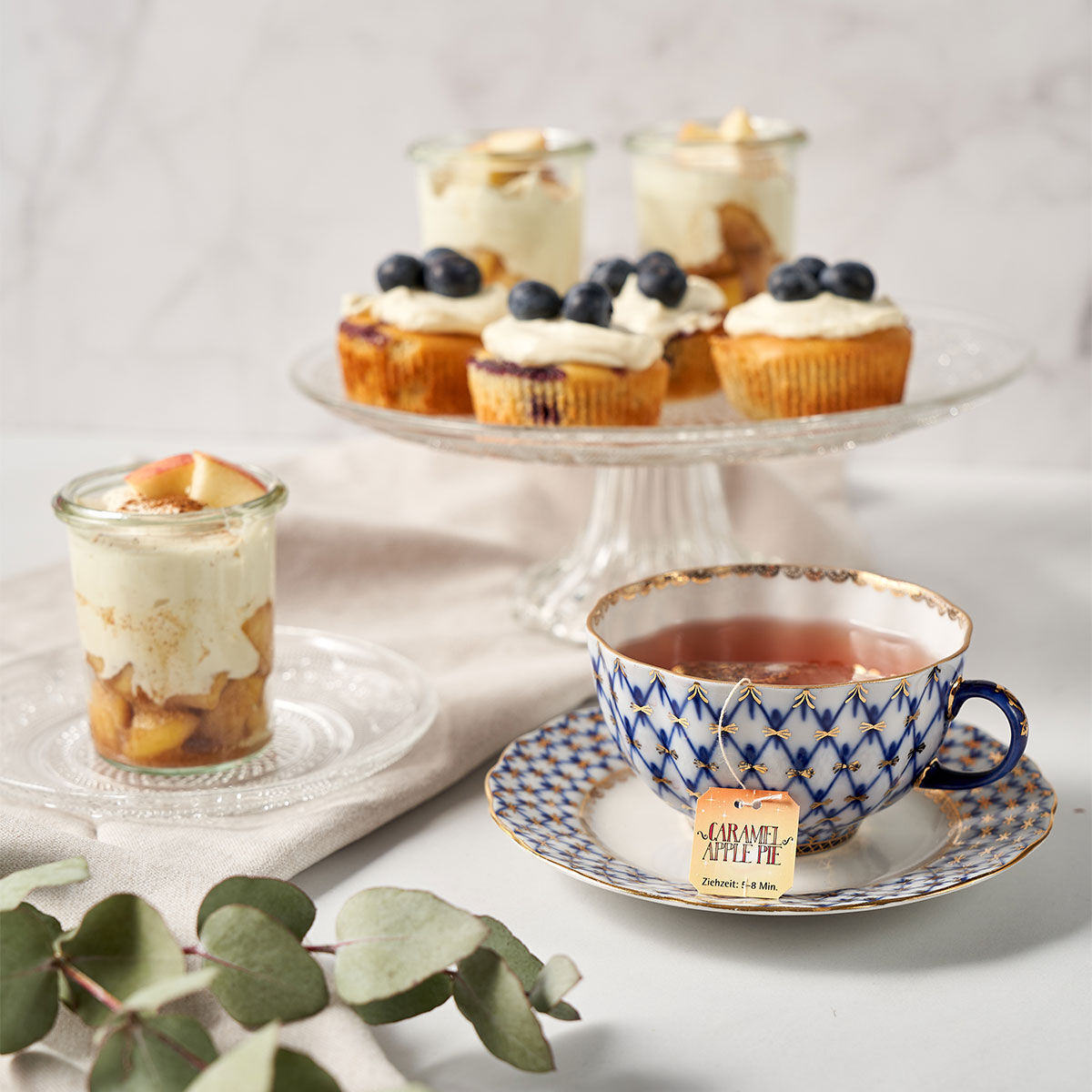 Eine Tasse Sweeteas Caramel Apple Pie-Tee steht neben Parfaits in Gläsern und Cupcakes mit Sahne und Blaubeeren, alles präsentiert auf einem Leuchttisch mit Eukalyptusblättern.