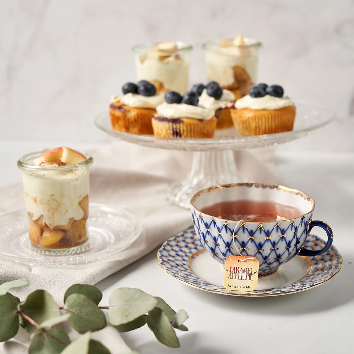 Eine Tasse Sweeteas Caramel Apple Pie-Tee steht neben Parfaits in Gläsern und Cupcakes mit Sahne und Blaubeeren, alles präsentiert auf einem Leuchttisch mit Eukalyptusblättern.