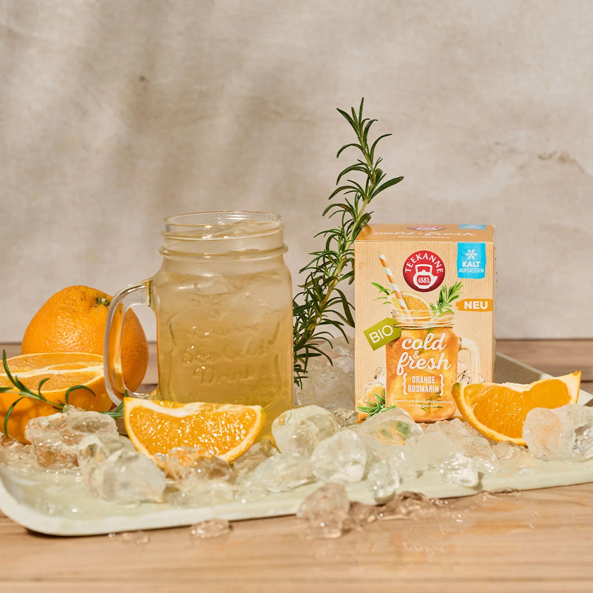 Ein Einmachglas mit Eistee steht auf einem Tablett mit Eiswürfeln, Orangenscheiben und Rosmarinzweigen. Dahinter befindet sich eine Verpackung mit der Aufschrift „Kalt & Frisch Orange-Rosmarin“, auf der Orangen und Symbole für Erfrischungsgetränke abgebil