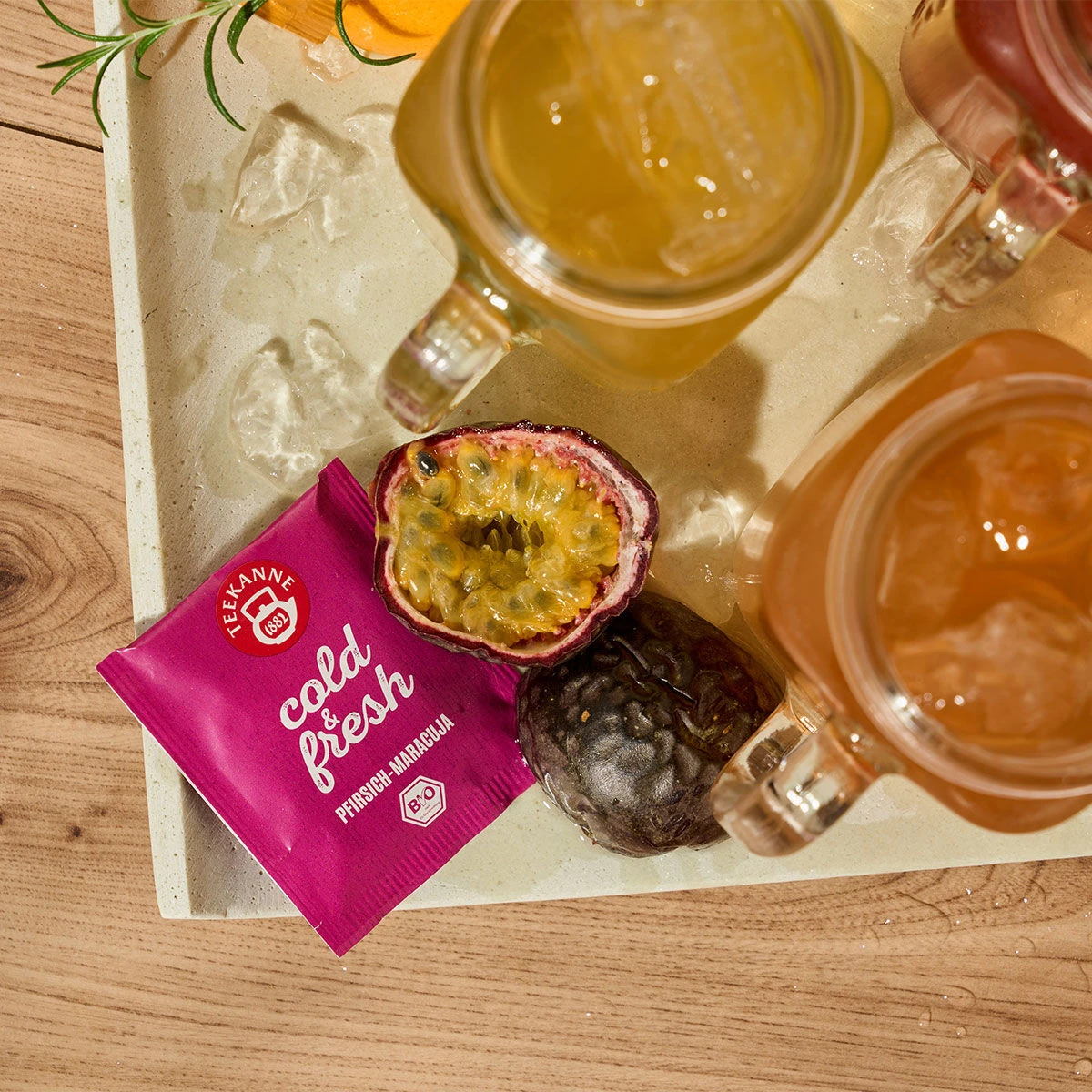 Eine aufgeschnittene Passionsfrucht, eine ganze Passionsfrucht und eine Packung Teekanne kalt & frisch Pfirsich-Maracuja stehen auf einem Tablett neben Einmachglasbechern, die mit eisgekühltem Erfrischungsgetränk gefüllt sind.