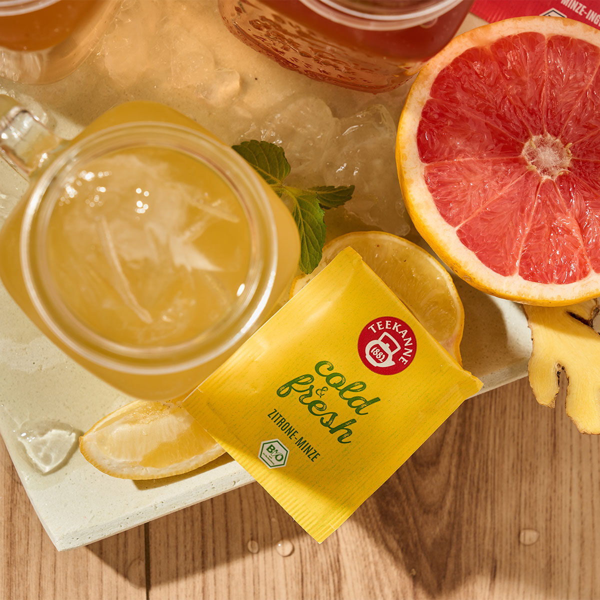 Ein Glas Eistee mit Zitronenscheiben, ein gelbes Teekanne Cold & Fresh Zitronen-Minze-Päckchen, eine halbe Grapefruit, Ingwerstücke und Getränke auf Eis – alles auf einer Holzoberfläche angerichtet für die ultimative Erfrischung.