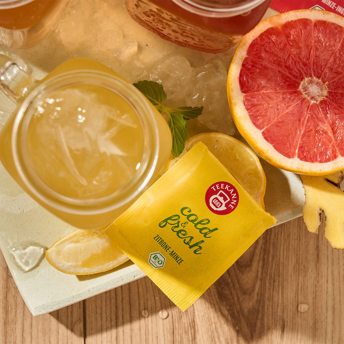 Ein Glas Eistee mit Zitronenscheiben, ein gelbes Teekanne Cold & Fresh Zitronen-Minze-Päckchen, eine halbe Grapefruit, Ingwerstücke und Getränke auf Eis – alles auf einer Holzoberfläche angerichtet für die ultimative Erfrischung.