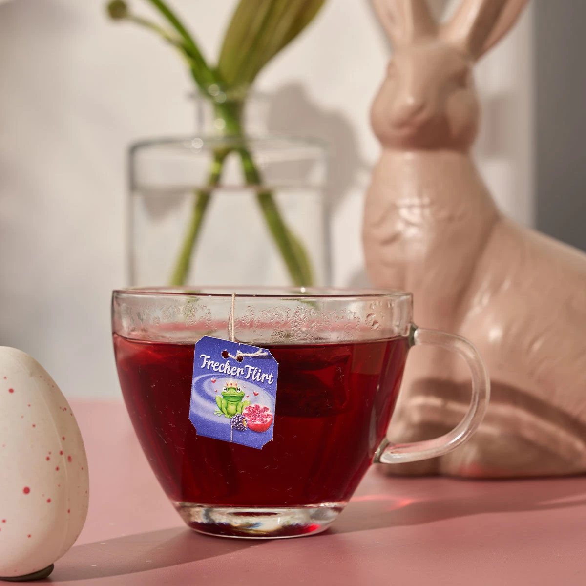 Eine klare Glastasse mit rotem Früchtetee und der Aufschrift „Frecher Flirt“ steht auf einem rosa Untergrund. Im Hintergrund sind ein Keramikhase, eine Vase mit Blumen und ein weiß gesprenkeltes Ei zu sehen.