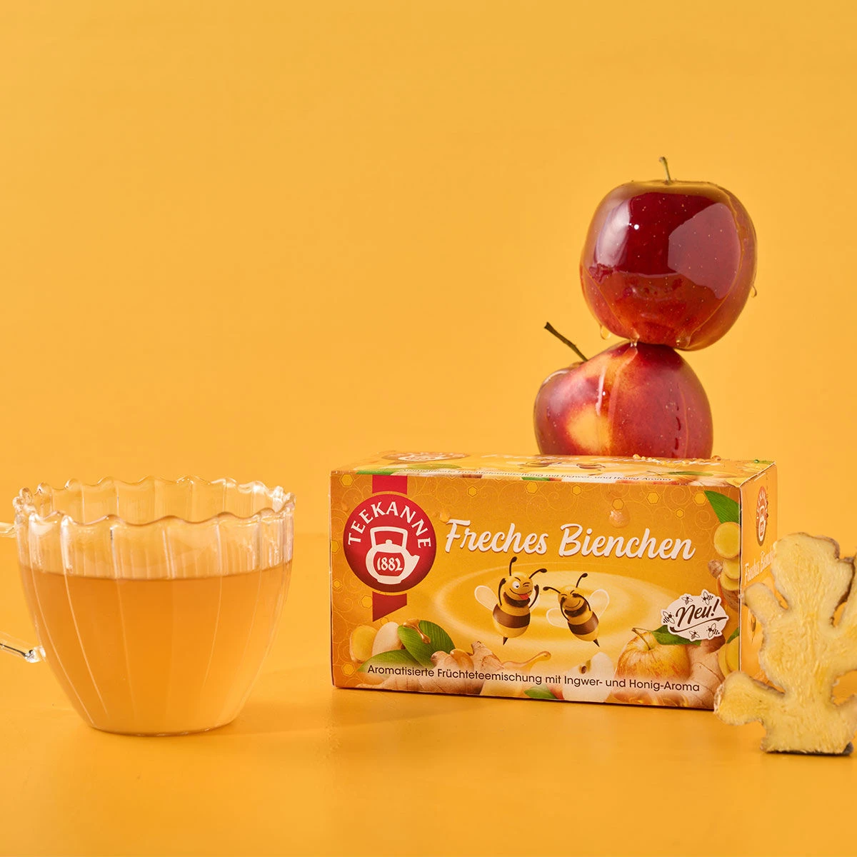 Auf einer gelben Fläche mit gelbem Hintergrund werden eine Glastasse Freches Bienchen-Tee, eine Schachtel Freches Bienchen, zwei gestapelte rote Äpfel und eine Scheibe Ingwer präsentiert.