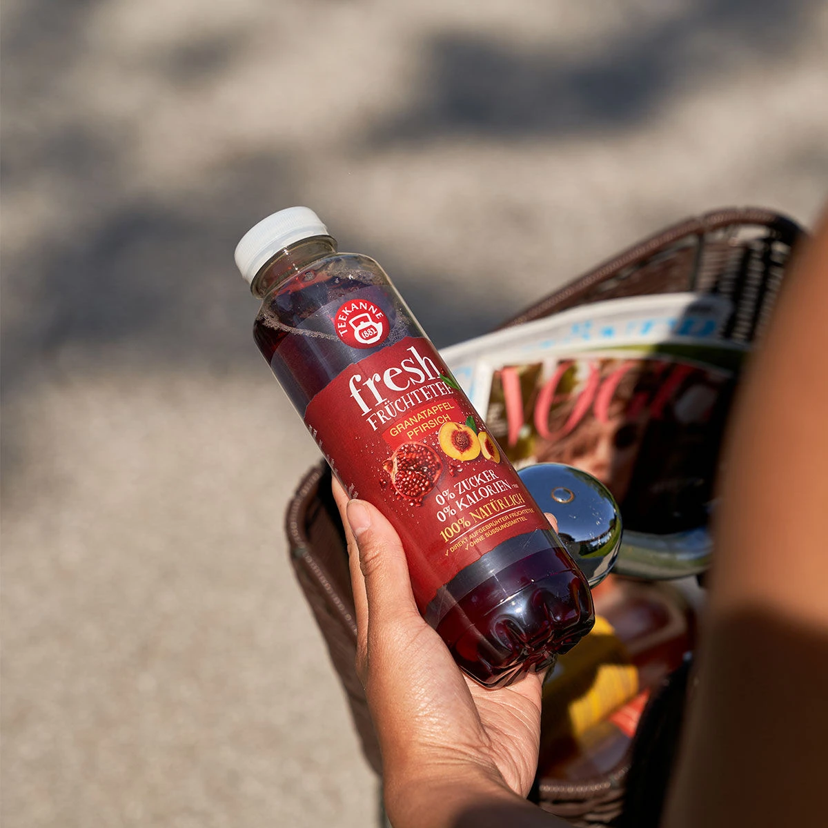 Eine Person hält eine Flasche aus dem Sixpack Fresh Granatapfel-Pfirsich über einen Korb voller Zeitschriften und anderer Artikel, wobei der Hintergrund leicht unscharf ist und eine Außenansicht zeigt.