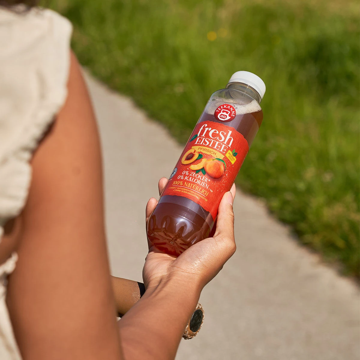 Jemand geht an einem sonnigen Tag im Freien spazieren und hält eine Flasche des Fresh Eistee Pfirsich Sixpack, einem Eistee mit Pfirsichgeschmack, in der Hand. Im Hintergrund sind verschwommenes Grün und ein Pfad zu sehen.