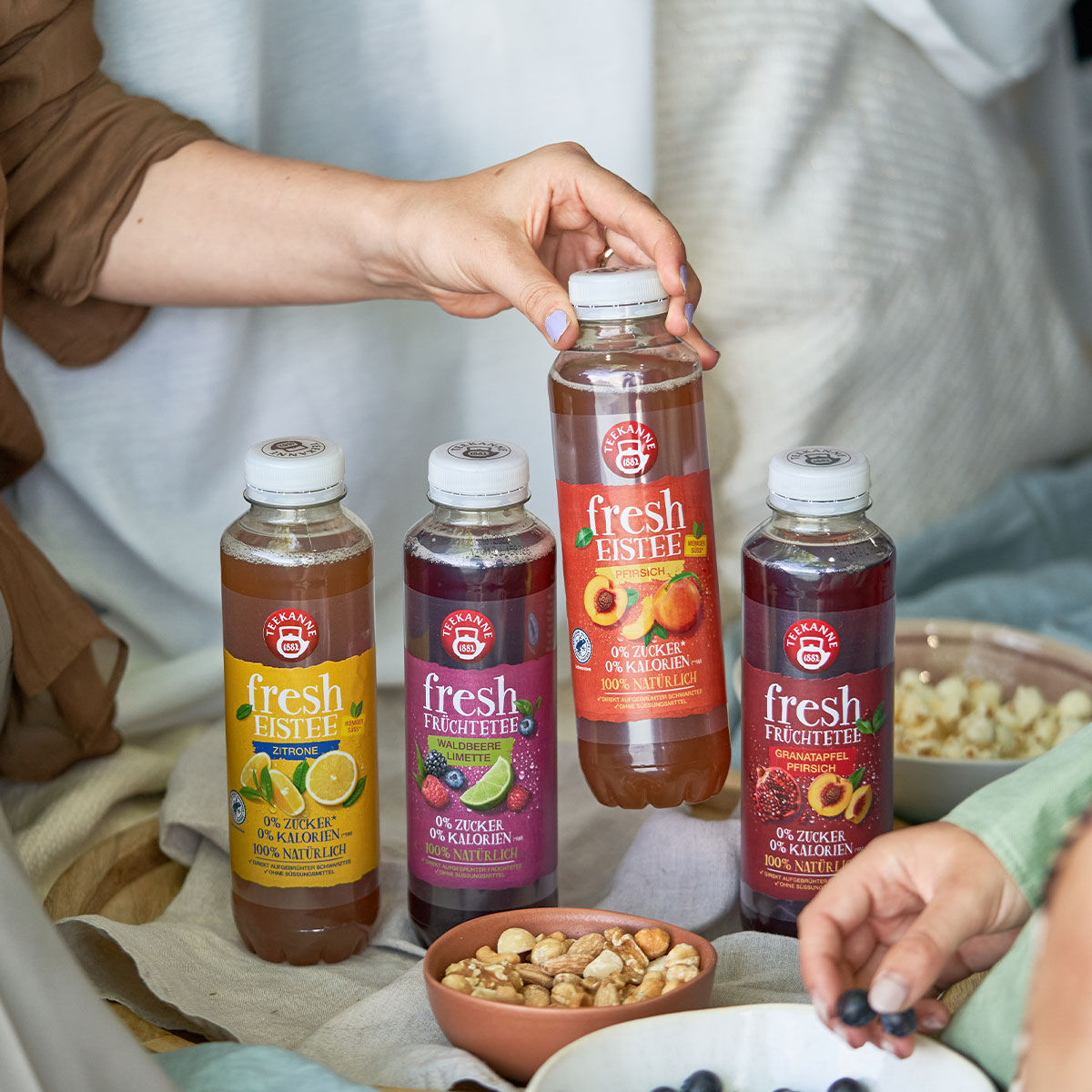 Vier Flaschen des frischen Probierset-Eistees stehen auf einem Tisch mit Snackschalen. Eine Person nimmt eine Flasche, eine andere greift nach einem Snack. Jede Flasche trägt ein individuelles Fruchtetikett für Abwechslung.