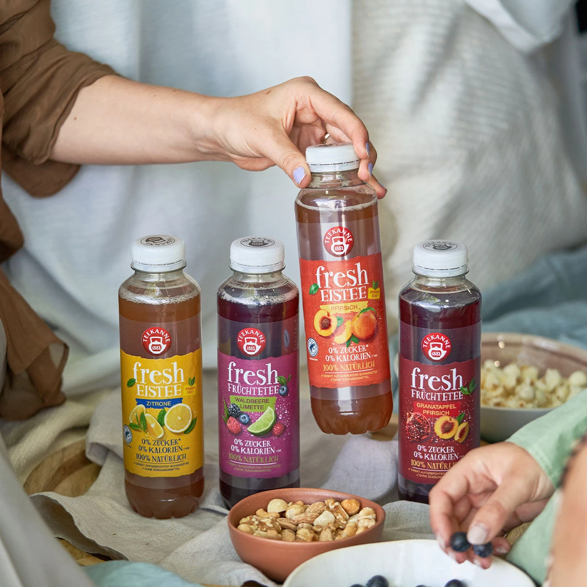 Vier Flaschen des frischen Probierset-Eistees stehen auf einem Tisch mit Snackschalen. Eine Person nimmt eine Flasche, eine andere greift nach einem Snack. Jede Flasche trägt ein individuelles Fruchtetikett für Abwechslung.