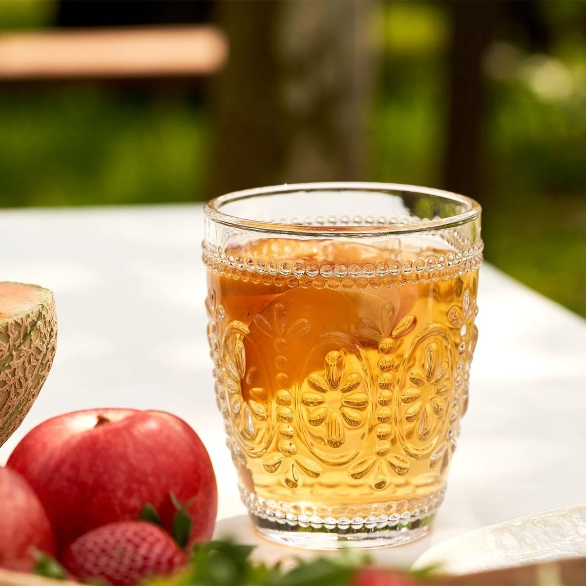 Ein Glas bernsteinfarbener Frio Classic Eistee Pfirsich steht auf einem weißen Tisch neben Äpfeln, Erdbeeren und Melonenscheiben, mit einem verzierten Glas und einem verschwommenen Außenhintergrund.