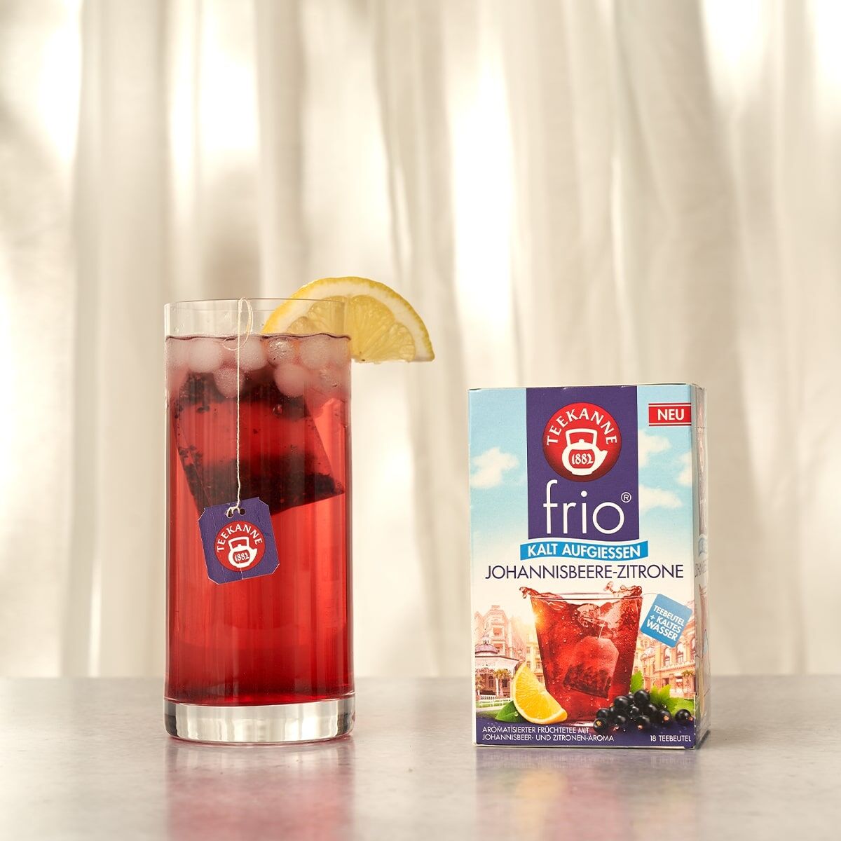 Ein Glas frío Johannisbeere-Zitrone, kalt serviert mit Zitronenscheibe und Teebeutel, steht neben einer Box frío Johannisbeere-Zitrone vor einem hellen Vorhang-Hintergrund.