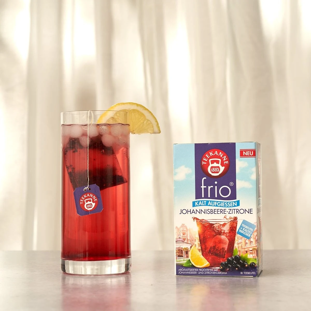 Ein Glas frío Johannisbeere-Zitrone, kalt serviert mit Zitronenscheibe und Teebeutel, steht neben einer Box frío Johannisbeere-Zitrone vor einem hellen Vorhang-Hintergrund.