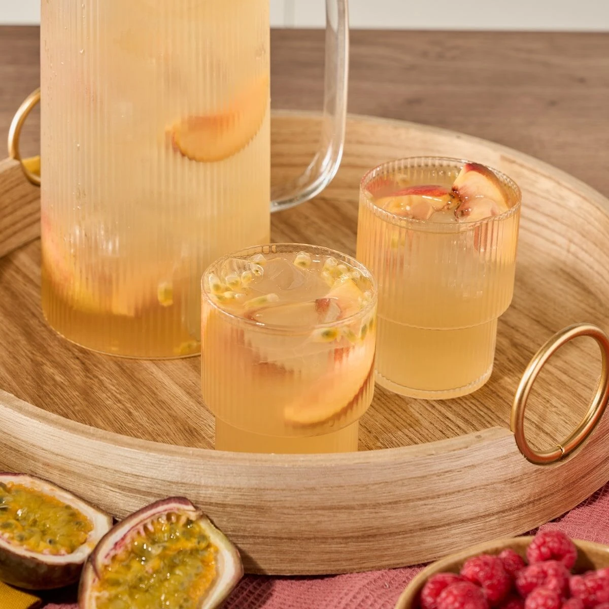 Ein Glaskrug und zwei geriffelte Gläser, gefüllt mit hellgelber Pfirsich-Maracuja, stehen auf einem Holztablett. Halbierte Maracuja und eine Schale Himbeeren verleihen diesem Erfrischungsgetränk einen farbenfrohen Touch.