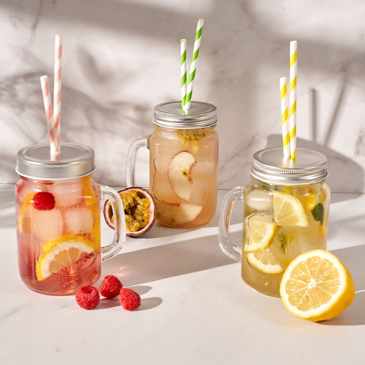 Drei Getränke im Einmachglas mit gestreiften Strohhalmen – eines mit Himbeeren und Zitrone, eines mit Pfirsichen und eines mit Zitrone und Minze – stehen neben einer halbierten Passionsfrucht und Himbeeren, ideal zum Genießen des erfrischenden Frio Probie