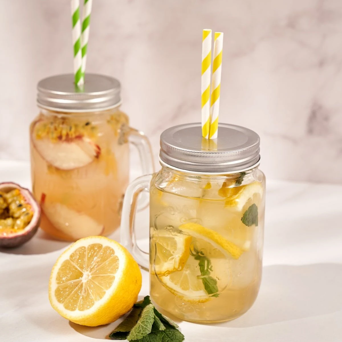 Zwei Einmachgläser mit Limonade, Zitronenscheiben, Minze und gestreiften Papierstrohhalmen stehen auf einer hellen Fläche. Eine halbierte Zitrone und Minzblätter sorgen für Frische, während Passionsfrucht- und Pfirsichscheiben das frio Sport Probierset he