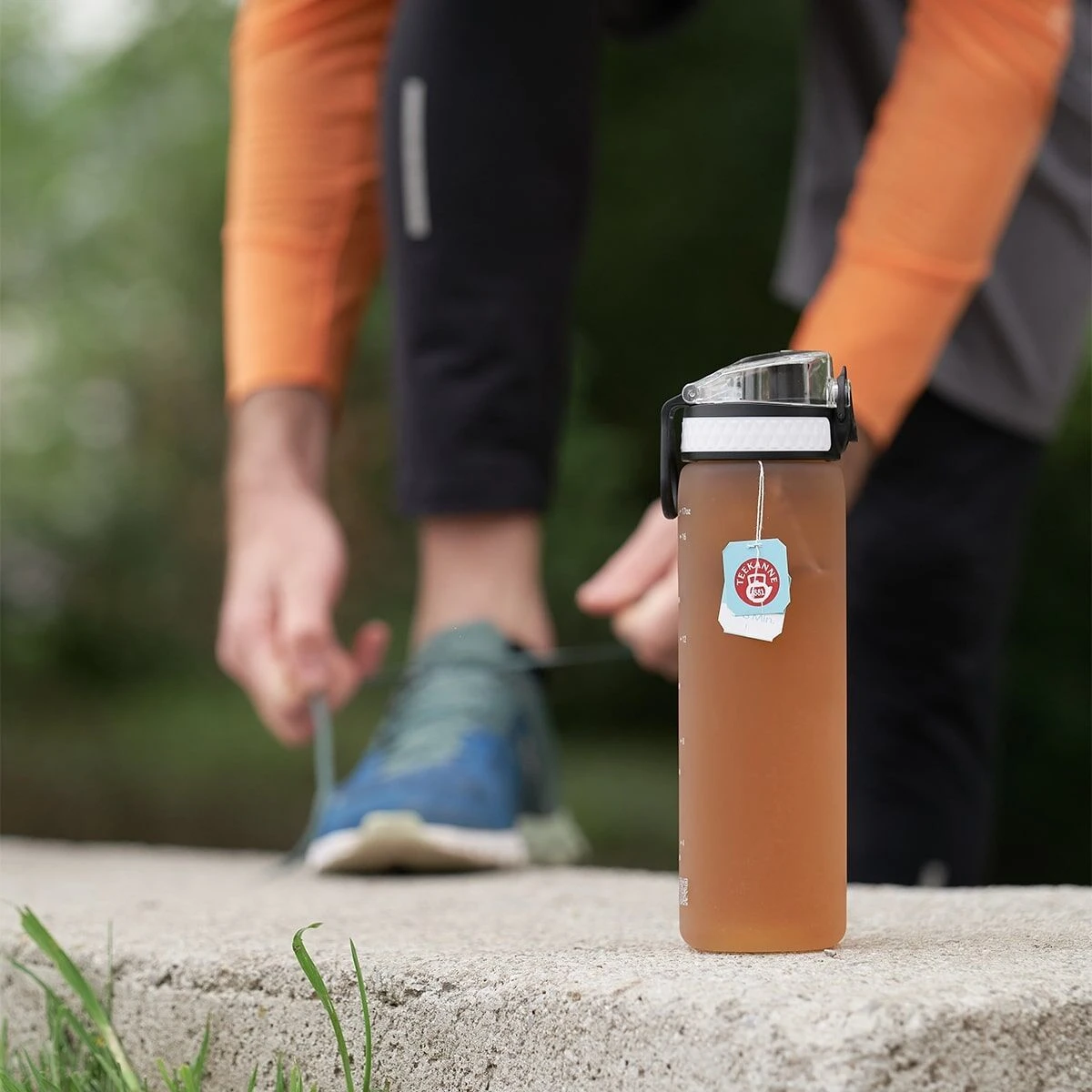 Eine Nahaufnahme einer mit frio Sport Aktiv Apfel-Zitrone gefüllten Wasserflasche steht auf einem Betonvorsprung, während im unscharfen Hintergrund eine Person in Sportkleidung blaue Laufschuhe schnürt. Draußen ist üppiges Grün zu sehen.
