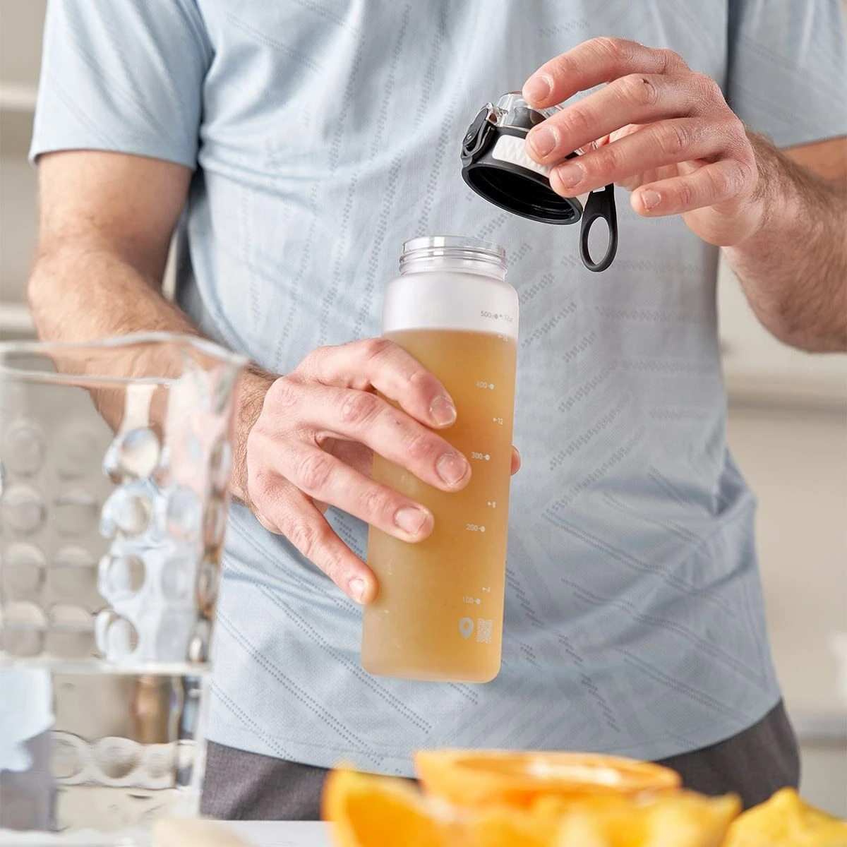 Eine Person im hellblauen Hemd öffnet eine Bügelflasche frio Sport Vital Mango-Orange, auf dem Tisch stehen ein Glas Wasser und Orangenscheiben, die an die erfrischenden Mango-Orangen-Aromen dieses Erfrischungsgetränks erinnern.
