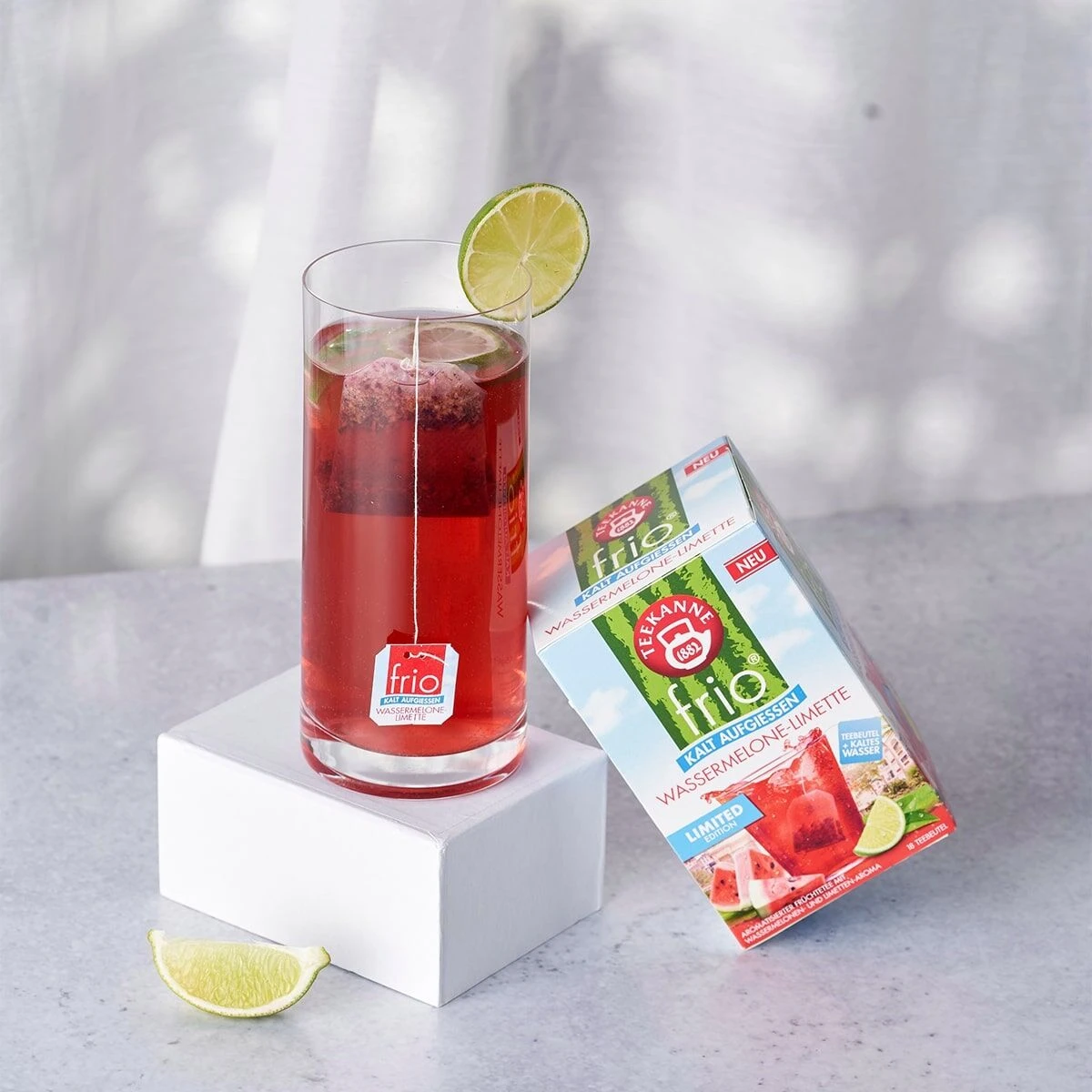 Ein hohes Glas roter Früchtetee mit Limette steht auf einem weißen Block. Daneben eine Packung Teekanne frio Wassermelone-Limette, ideal als Kaltaufguss-Tee. Im Hintergrund ist eine weiße Gardine zu sehen.