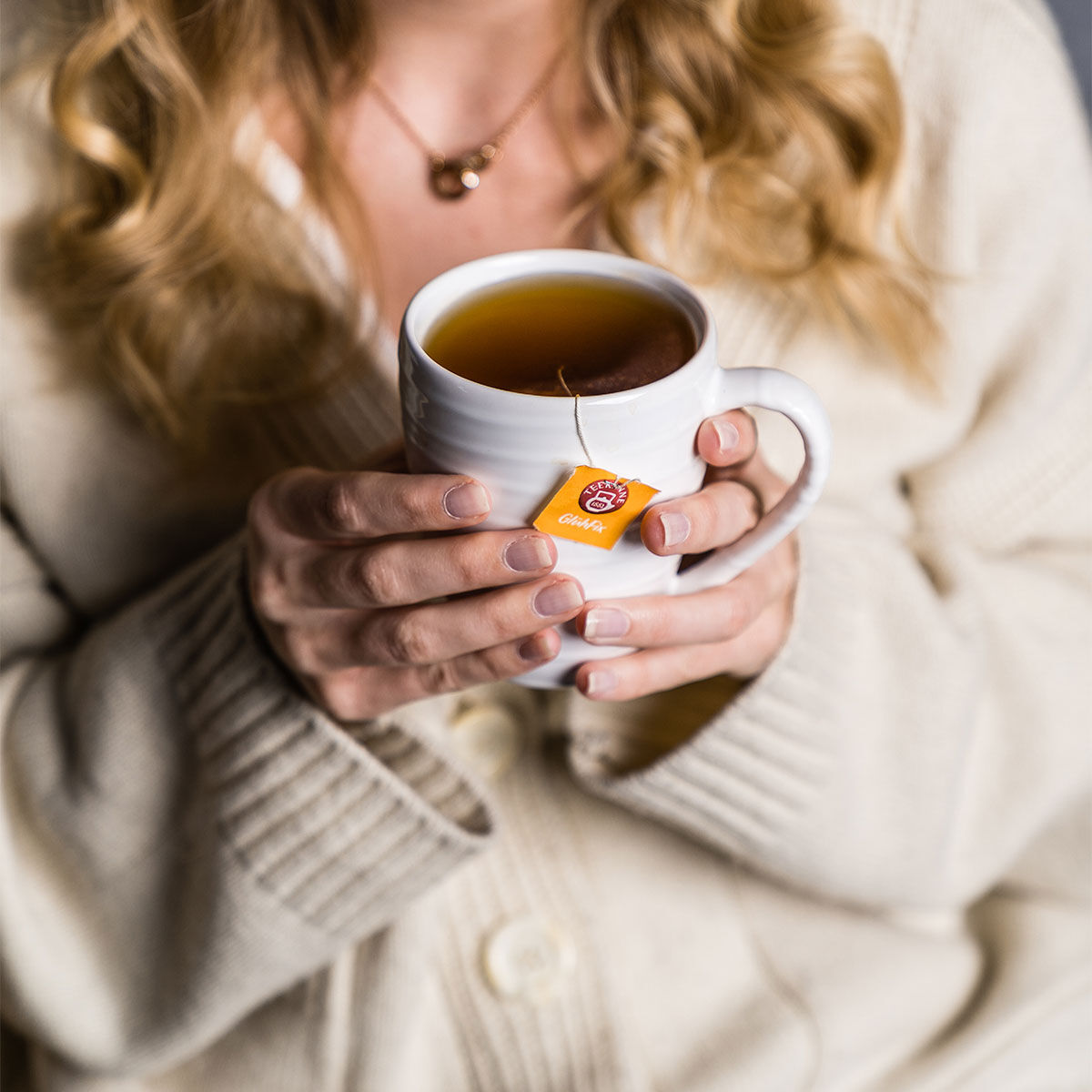 Eine Frau mit welligem blondem Haar und Naturnägeln hält eine weiße Tasse Tee mit der Aufschrift „Glühfix“. Sie trägt einen cremefarbenen Pullover. Nur ihre Hände, ein Teil ihres Gesichts und ihr Oberkörper sind zu sehen.