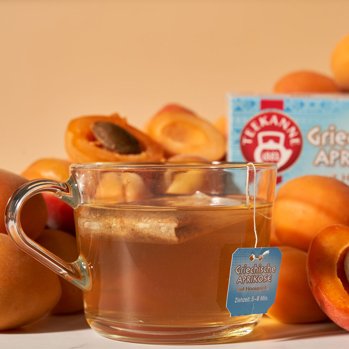 Eine Tasse heißen griechischen Aprikosentees mit einem Teebeutel steht vor frischen Aprikosen und der Produktverpackung, alles warm beleuchtet vor einem pfirsichfarbenen Hintergrund.