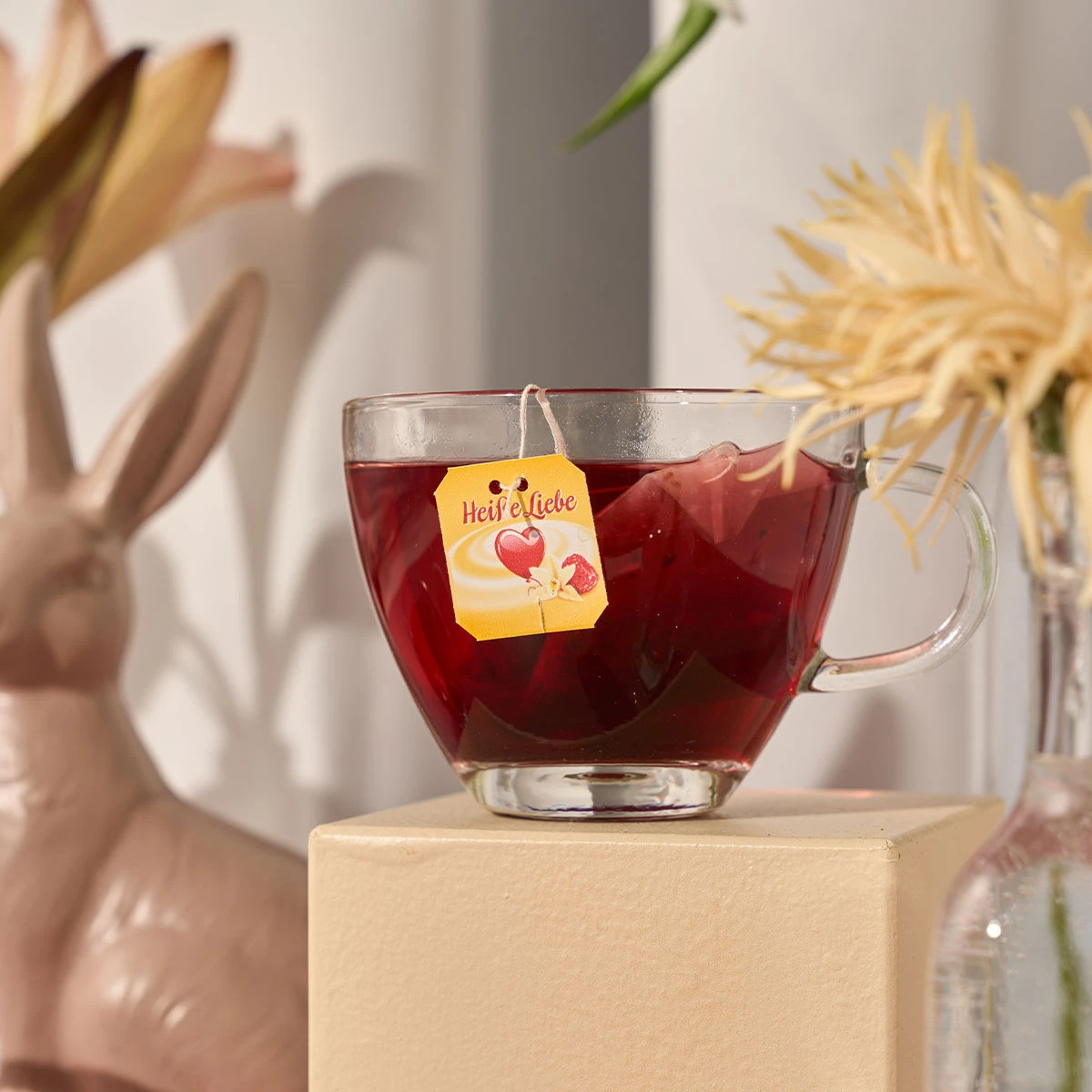 Eine klare Glastasse mit Himbeer-Rottee von Heiße Liebe steht auf einem beigen Sockel. Ein gelber Heiße Liebe-Teebeutel hängt über dem Rand. Im Hintergrund sind ein Keramikhase, Blumen und eine Glasvase zu sehen.