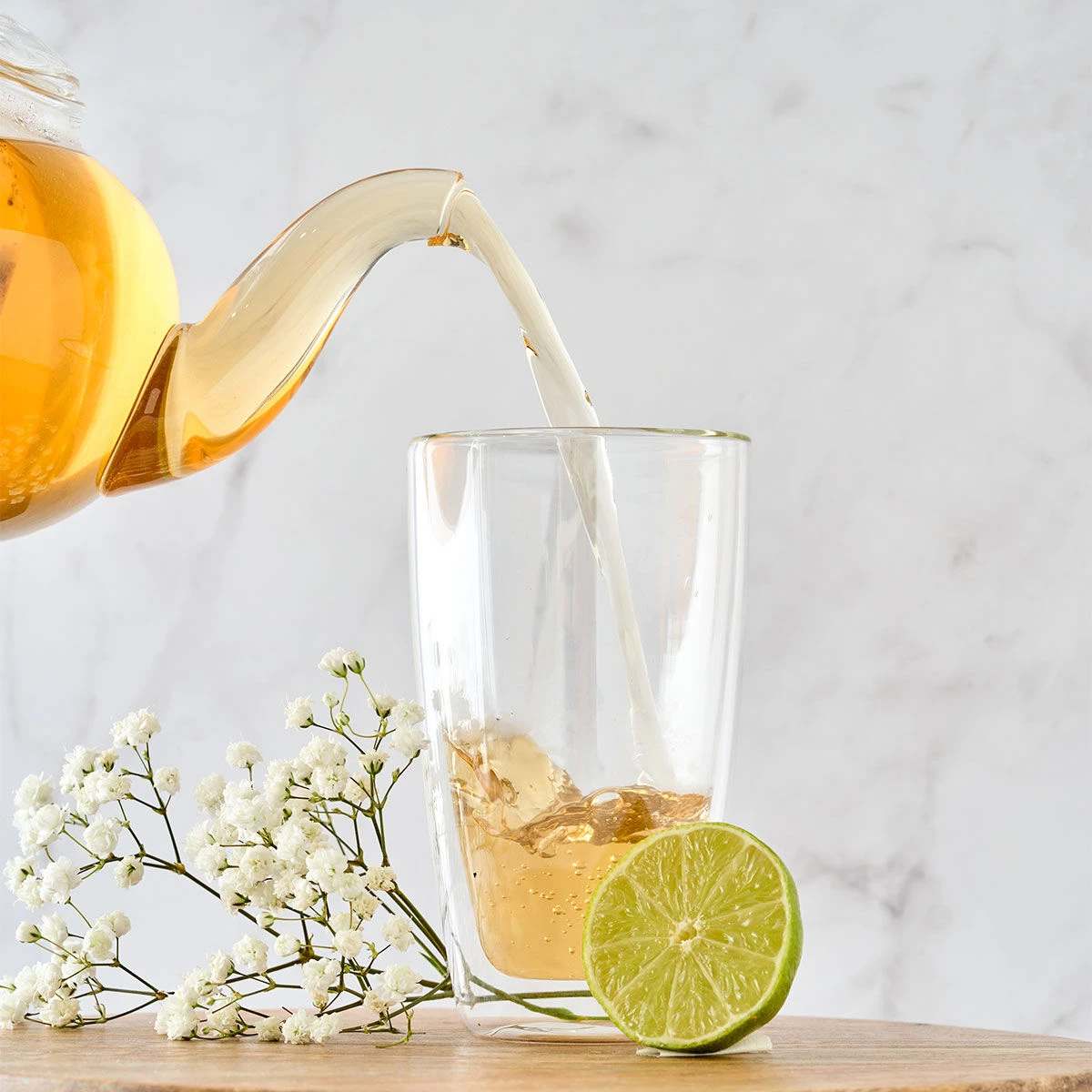 Aus einer Glasteekanne wird Heißer Hugo in ein klares Glas auf einer Holzoberfläche gegossen. Daneben stehen frische Limettenhälften und weiße Holunderblüten, alles vor einem hellen Marmorhintergrund.