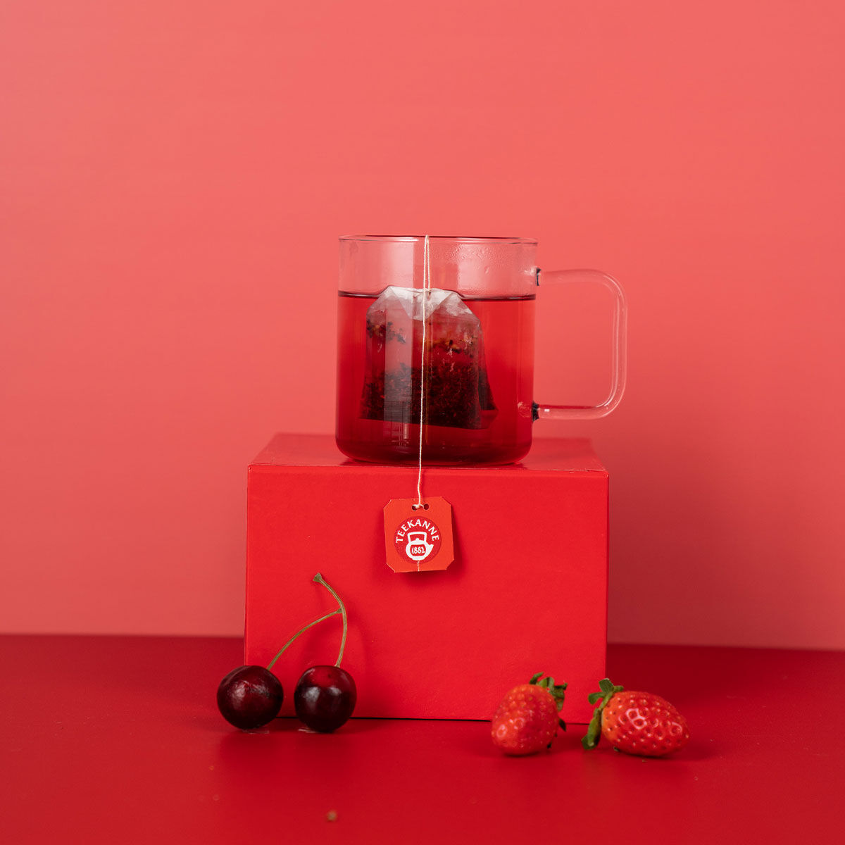Eine Glastasse mit rotem Tee und einem Teebeutel steht auf einer roten Schachtel neben Herzkirschen und Erdbeeren vor einem leuchtend roten Hintergrund.