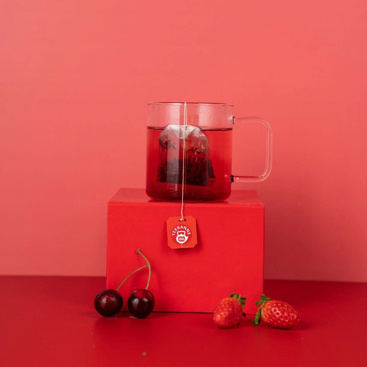 Eine Glastasse mit rotem Tee und einem Teebeutel steht auf einer roten Schachtel neben Herzkirschen und Erdbeeren vor einem leuchtend roten Hintergrund.