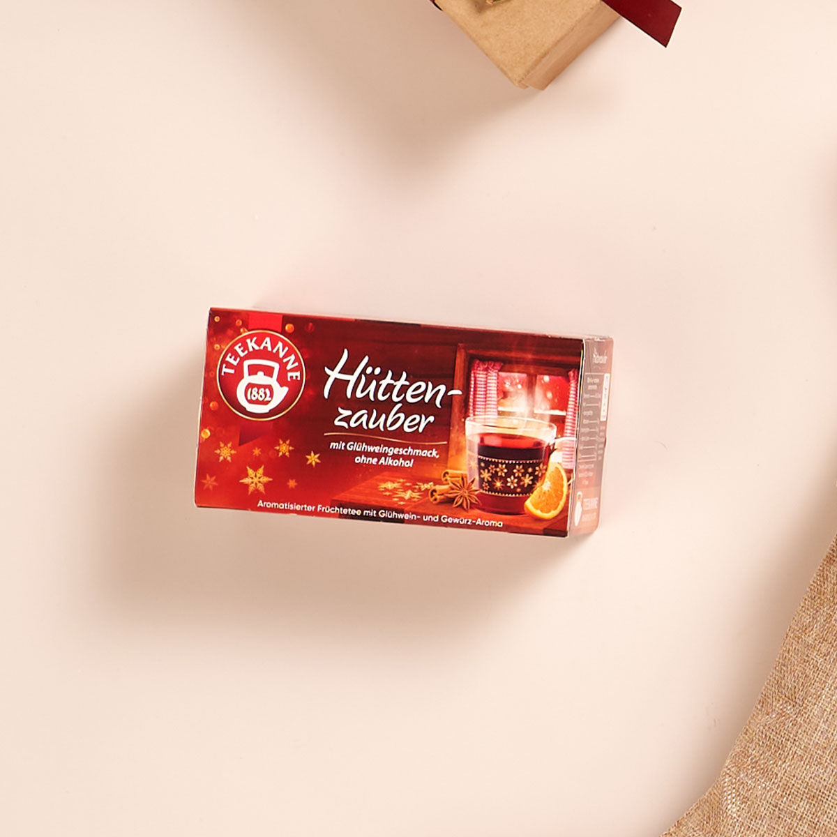 Auf einem hellen Untergrund steht eine rote Schachtel Teekanne Hüttenzauber. Darin ist eine dampfende Tasse mit Orangenscheiben und Gewürzen zu sehen. Der deutsche Text beschreibt ihn als fruchtig-würzigen Wintertee.