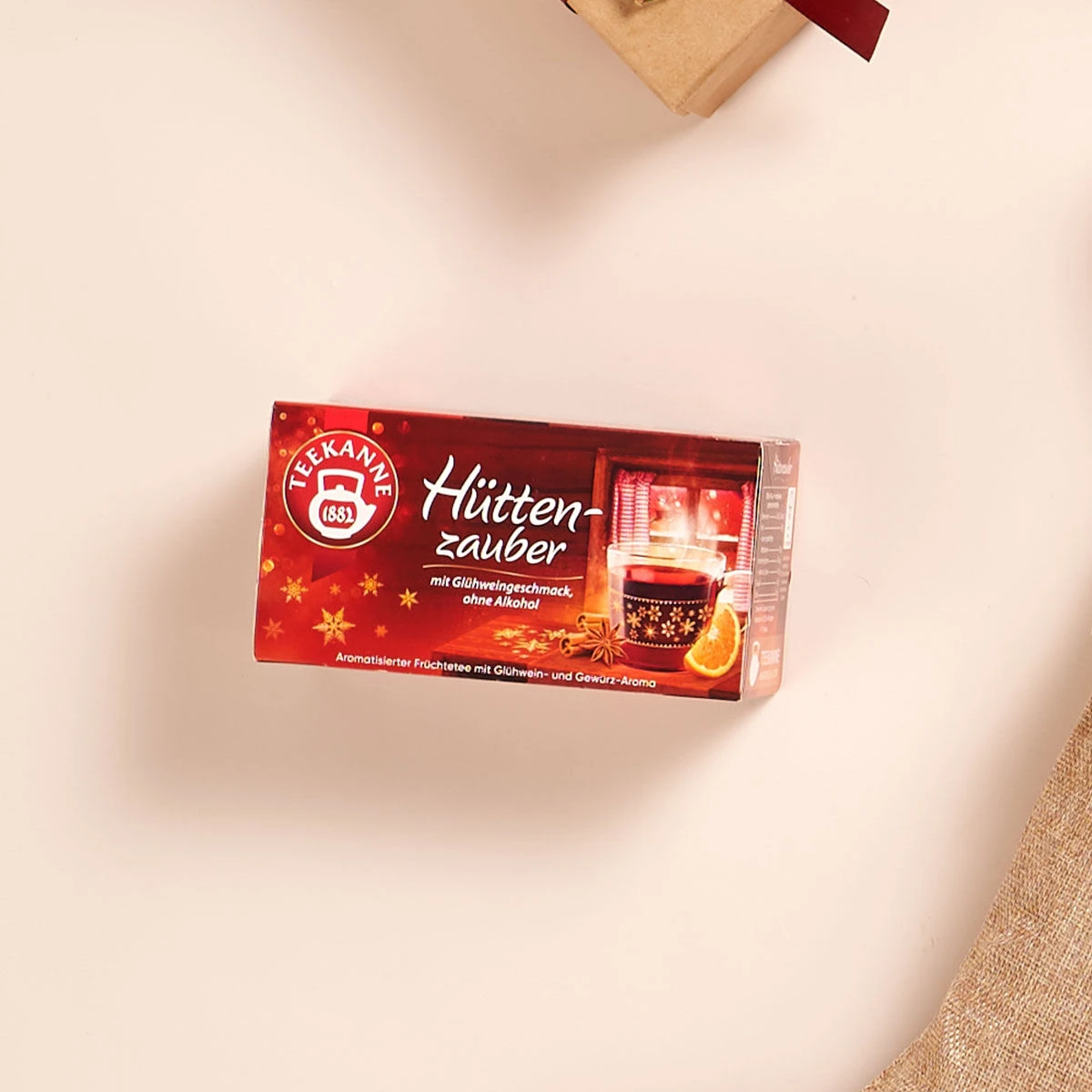 Auf einem hellen Untergrund steht eine rote Schachtel Teekanne Hüttenzauber. Darin ist eine dampfende Tasse mit Orangenscheiben und Gewürzen zu sehen. Der deutsche Text beschreibt ihn als fruchtig-würzigen Wintertee.