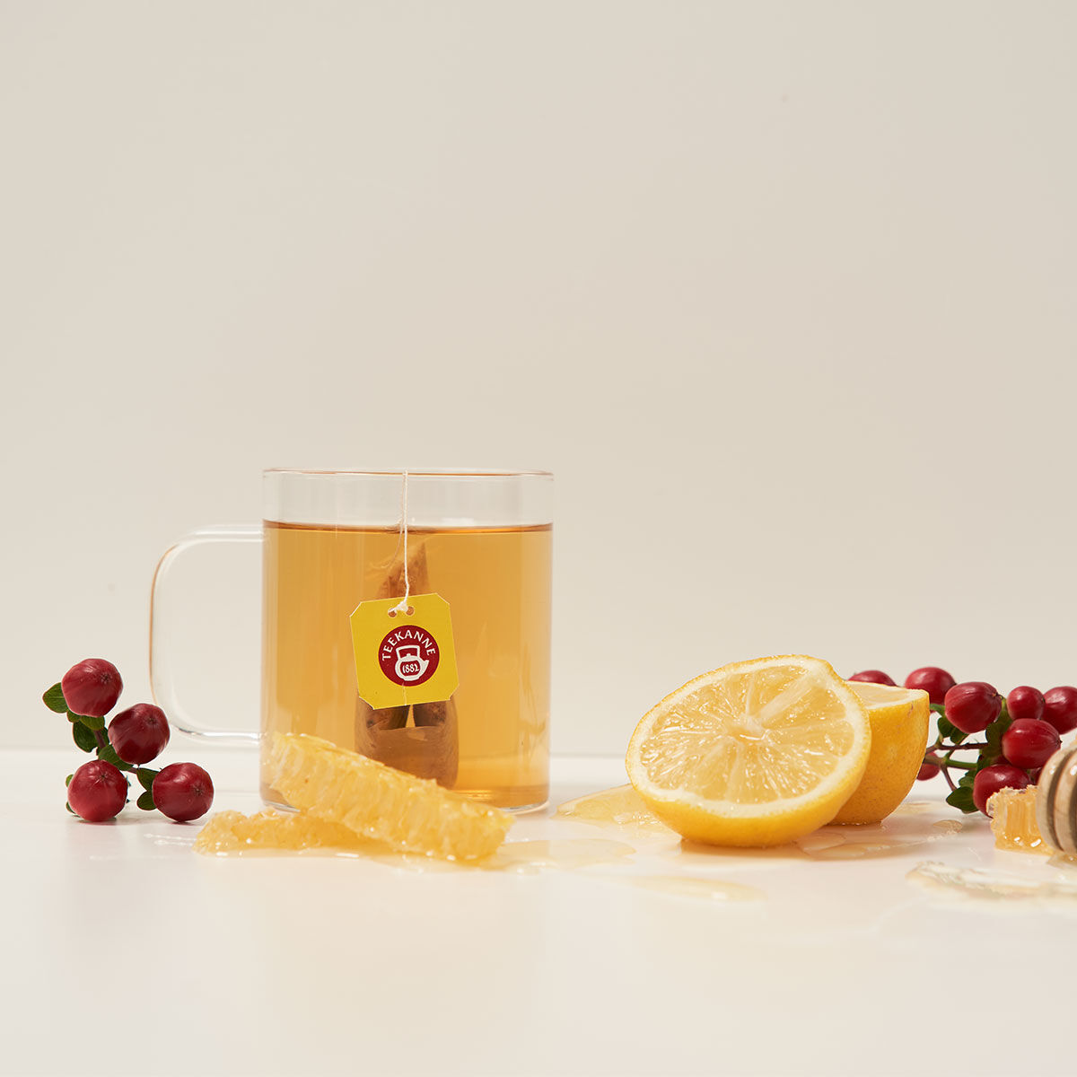 Eine klare Glastasse mit Italienischem Limone-Tee und einem Teebeutel, umgeben von Preiselbeeren, Zitronenscheiben, Honigwabe und Honig auf einer weißen Oberfläche mit hellem Hintergrund.
