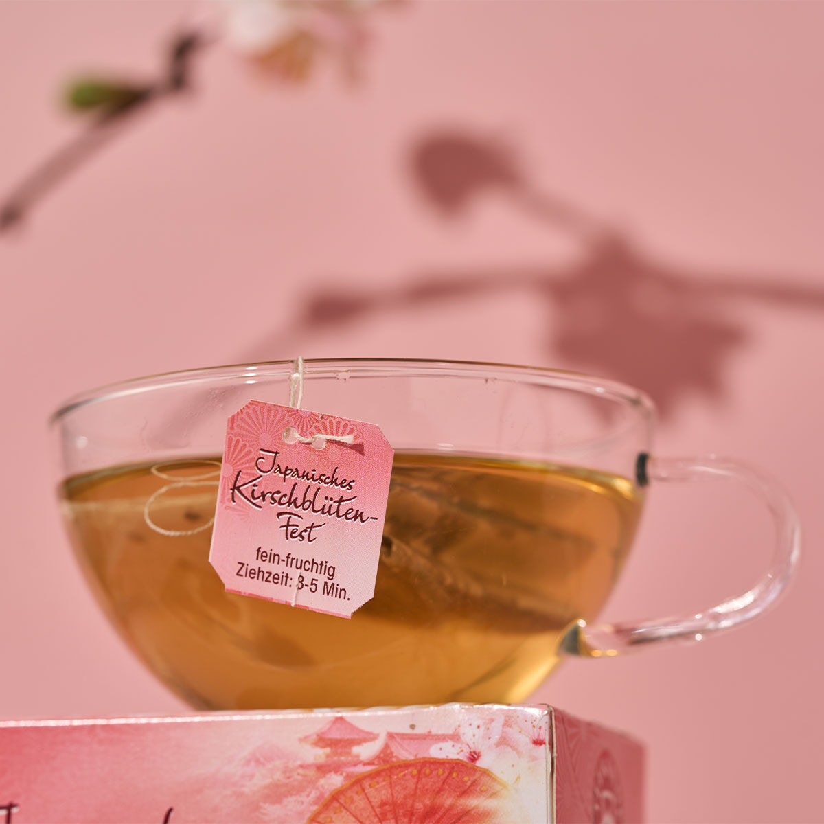 Eine Tasse Tee aus klarem Glas mit dem rosafarbenen Etikett „Japanisches Kirschblütenfest“ steht auf einer rosafarbenen Schachtel mit verschwommenen Kirschblüten und rosafarbenem Hintergrund und erinnert an den zarten Geschmack von „Weißem Tee mit Kirsche
