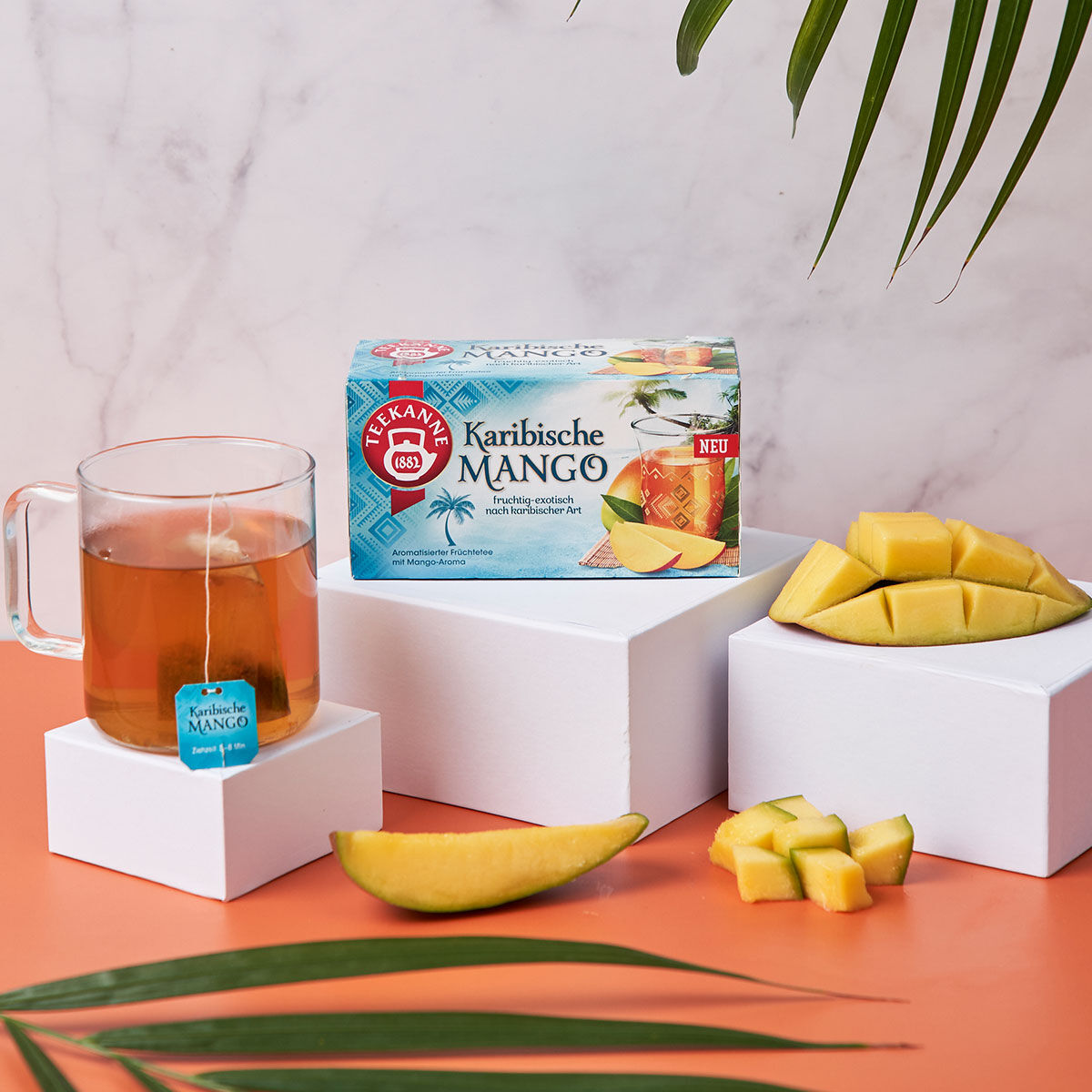 Eine Glastasse mit Karibische-Mango-Tee steht neben der Schachtel, umgeben von frischen Mangoscheiben auf weißen Blöcken mit Palmblättern darüber und einer orangefarbenen Oberfläche – ein perfektes tropisches Teebündel.