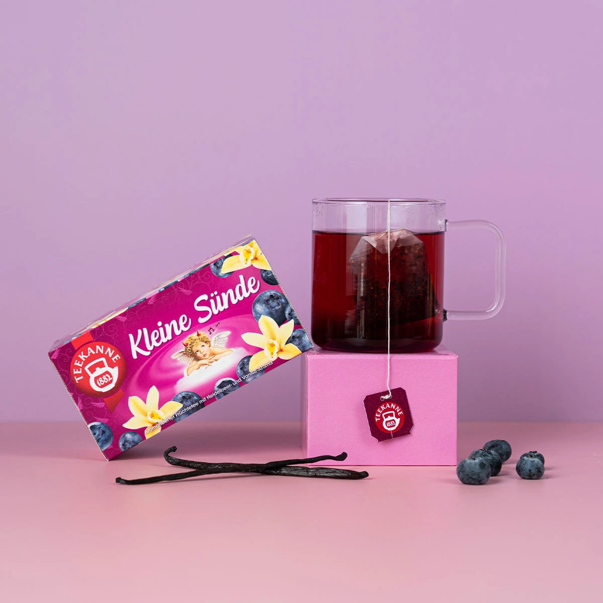Eine Schachtel Kleine Sünde-Tee mit Blaubeer- und Vanille-Motiven steht neben einer Glastasse mit rotem Tee auf einem rosa Würfel, umgeben von Blaubeeren und Vanilleschoten vor einem violetten Hintergrund.