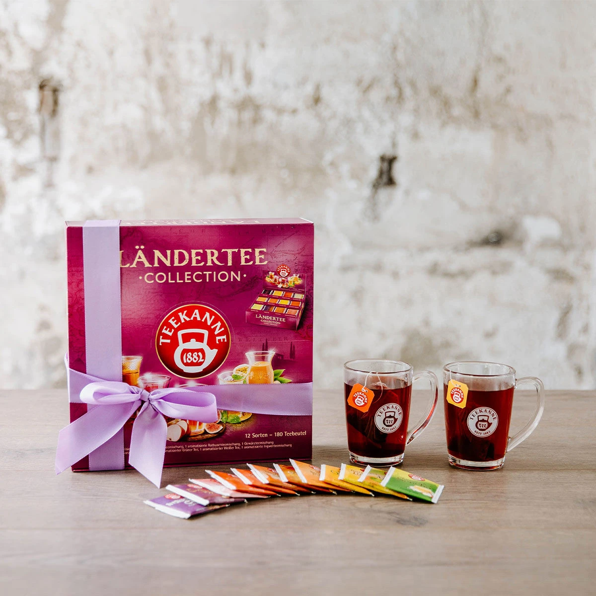 Eine Schachtel Ländertee-Collection Geschenkset mit 180 Beuteln, umwickelt mit einer lavendelfarbenen Schleife, steht auf einem Holztisch neben zwei Glasbechern und farbenfrohen Teebeuteln. Die rustikale, weiß getünchte Wand verleiht dieser einladenden Sz