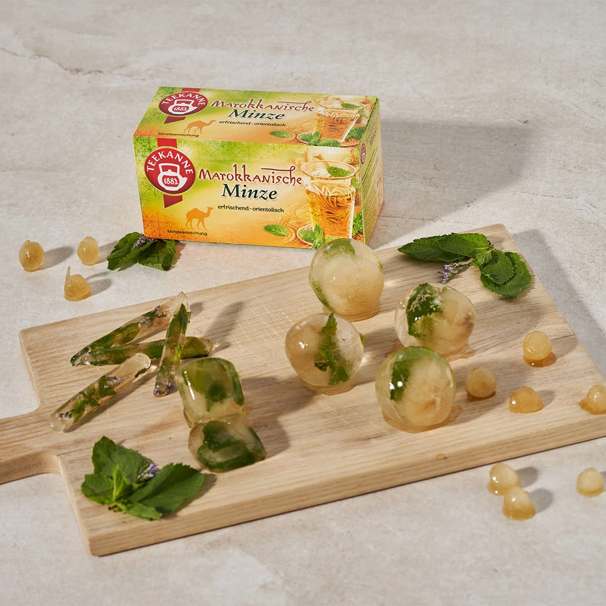 Eine Schachtel Marokkanischer Minze-Tee steht hinter einem Holzbrett, das mit runden und dreieckigen Eiswürfeln mit Minze bedeckt ist. Verstreute Minzblätter und glänzende Goldperlen runden die lebendige Szenerie der Minzteemischung ab.