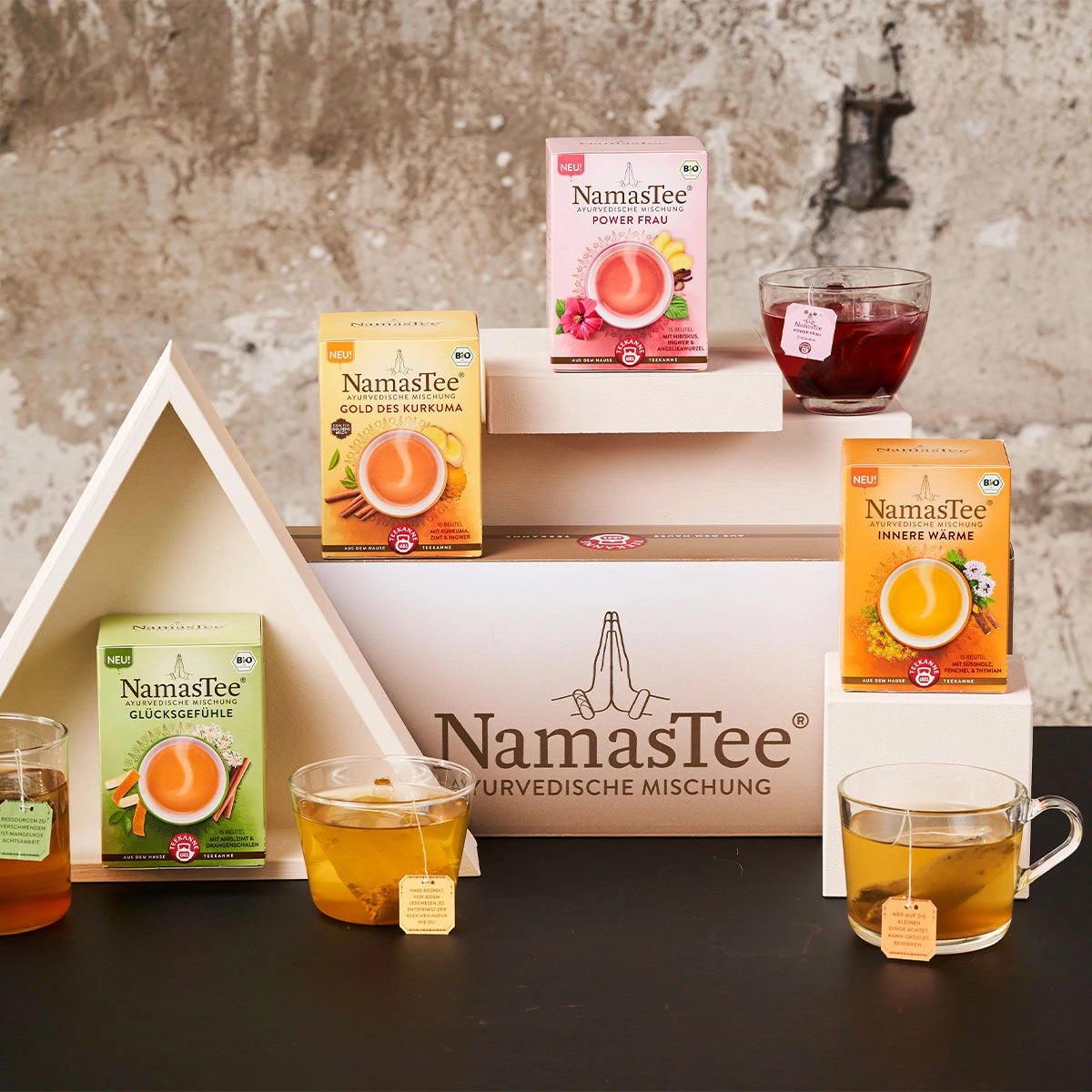 Eine Ausstellung von NamasTee® Achtsamkeitsset-Teeboxen mit Glastassen, angeordnet auf geometrischen weißen Ständern vor einer strukturierten Betonwand – ideal für Ihre achtsamen Momente.