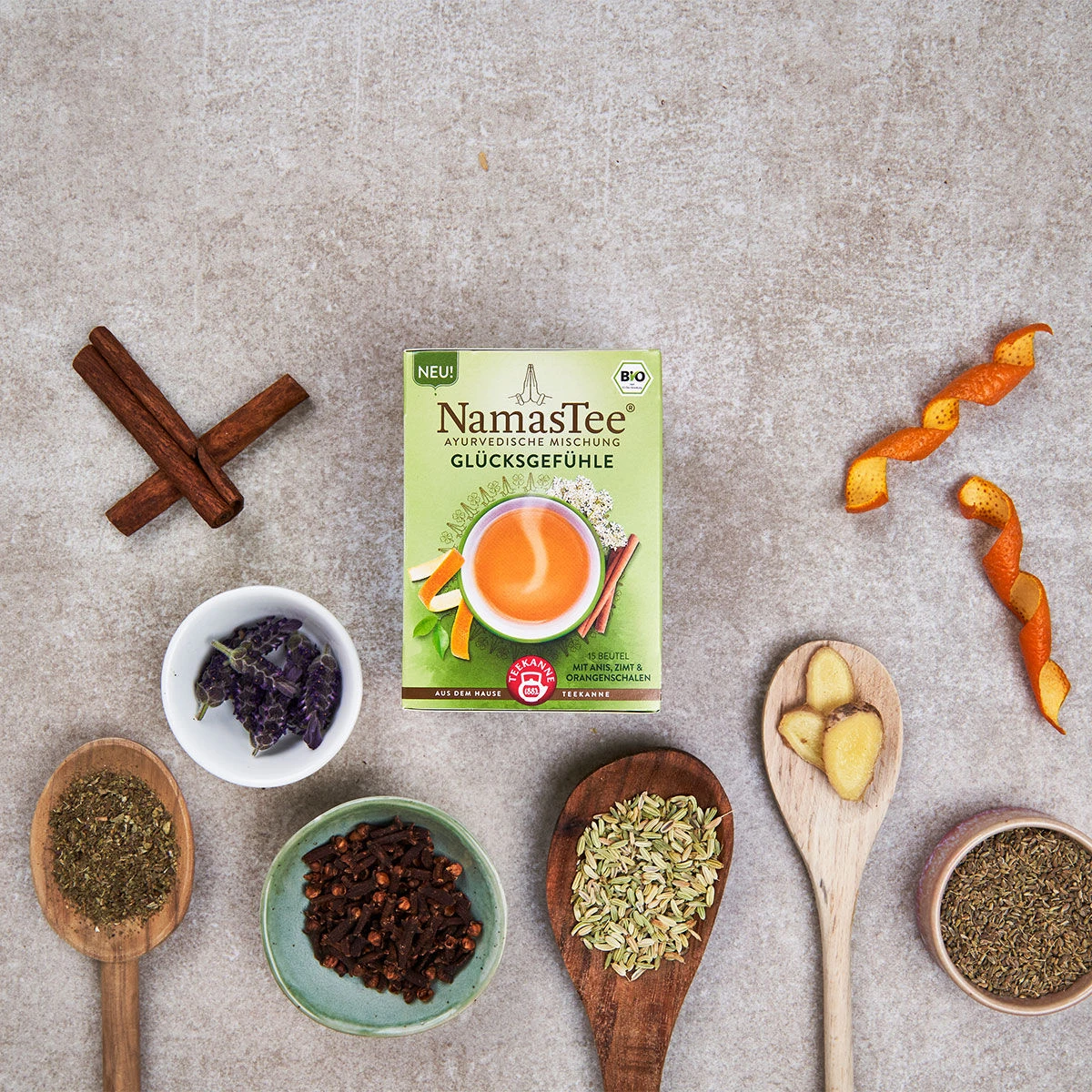 Eine Schachtel NamasTee® Glücksgefühle ist umgeben von Holzlöffeln mit Anis, Zimt, Ingwerscheiben sowie Schüsseln mit getrockneten Zutaten und Orangenschalen, alles hübsch auf einer Steinoberfläche angerichtet.