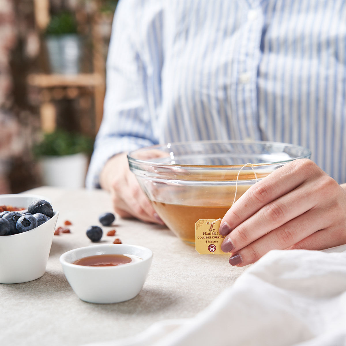 Eine Person im gestreiften Hemd hält einen NamasTee® Gold des Kurkuma Teebeutel über eine klare Glasschale mit Tee. Auf dem Tisch stehen Blaubeeren, Honig und Nüsse – die perfekte Beilage zum Gold des Kurkuma.