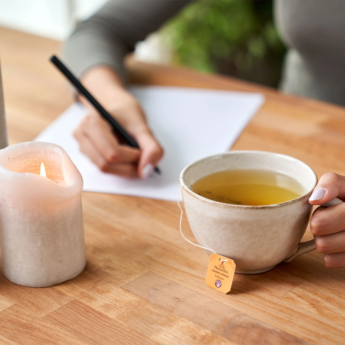 Eine Person schreibt auf einem Holztisch und hält eine Tasse NamasTee® Innere Wärme in der Hand, auf der das Etikett des Teebeutels zu sehen ist. In der Nähe steht eine teilweise geschmolzene weiße Kerze auf dem Tisch.