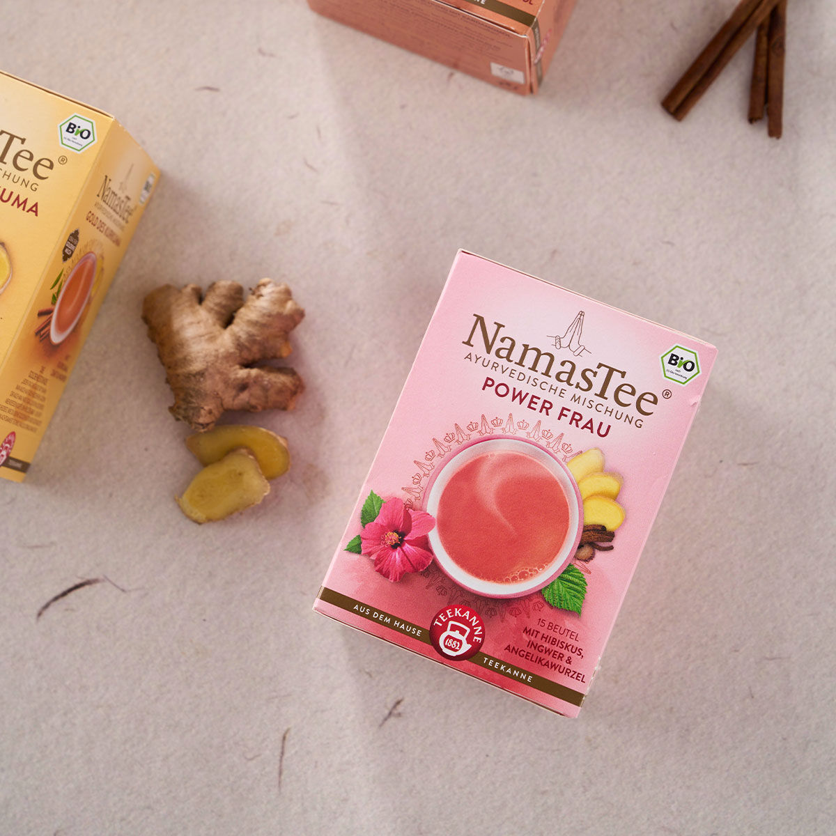 Eine NamasTee® Powerfrau Teedose steht auf einer hellen Fläche neben frischen Ingwerscheiben und einer weiteren Teedose. Die rosa Verpackung zeigt florale Grafiken und eine Teetasse. In der oberen rechten Ecke sind Zimtstangen zu sehen.