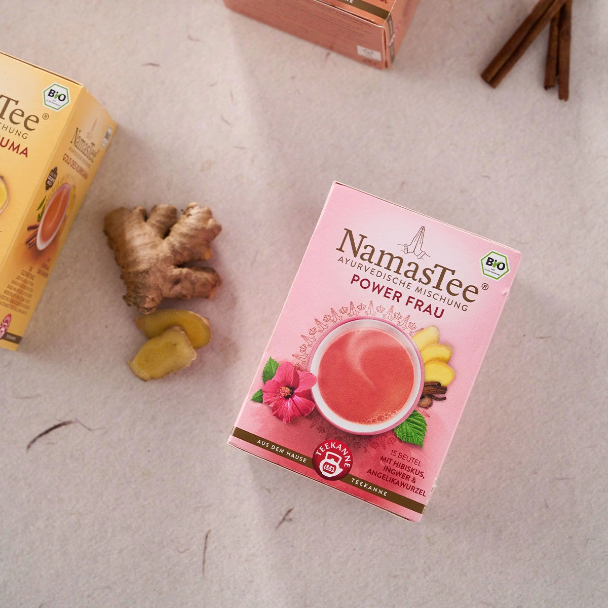Eine NamasTee® Powerfrau Teedose steht auf einer hellen Fläche neben frischen Ingwerscheiben und einer weiteren Teedose. Die rosa Verpackung zeigt florale Grafiken und eine Teetasse. In der oberen rechten Ecke sind Zimtstangen zu sehen.