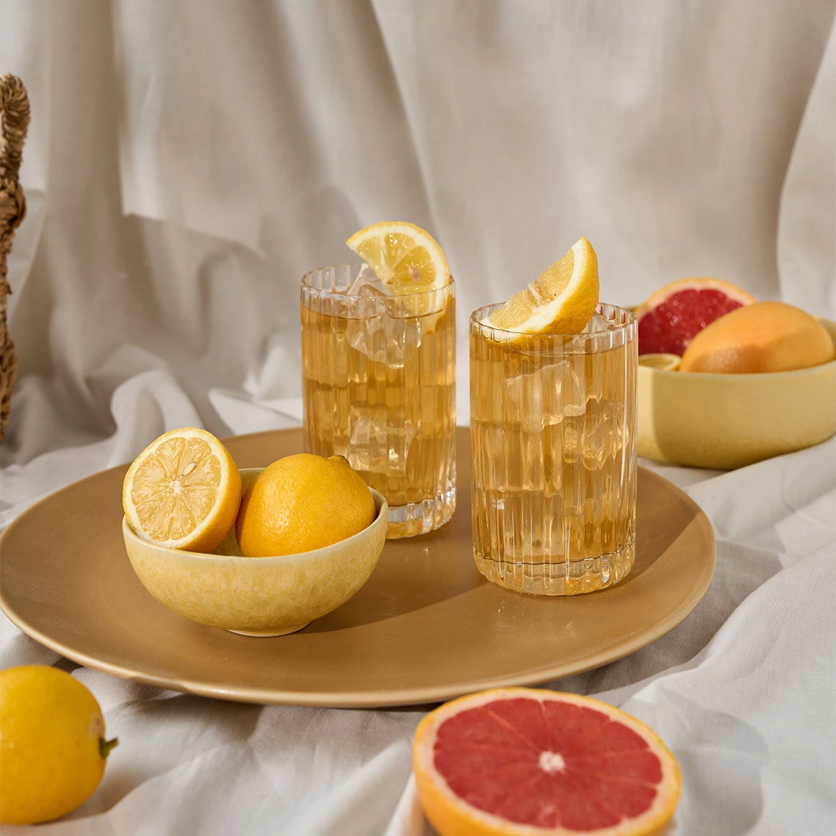 Zwei Gläser Eistee von ORGANICS Dankeschön stehen auf einem goldenen Teller mit einer Schale Zitronen. Grapefruitscheiben und ganze Zitrusfrüchte sind kunstvoll auf einem weißen Stoffhintergrund arrangiert.