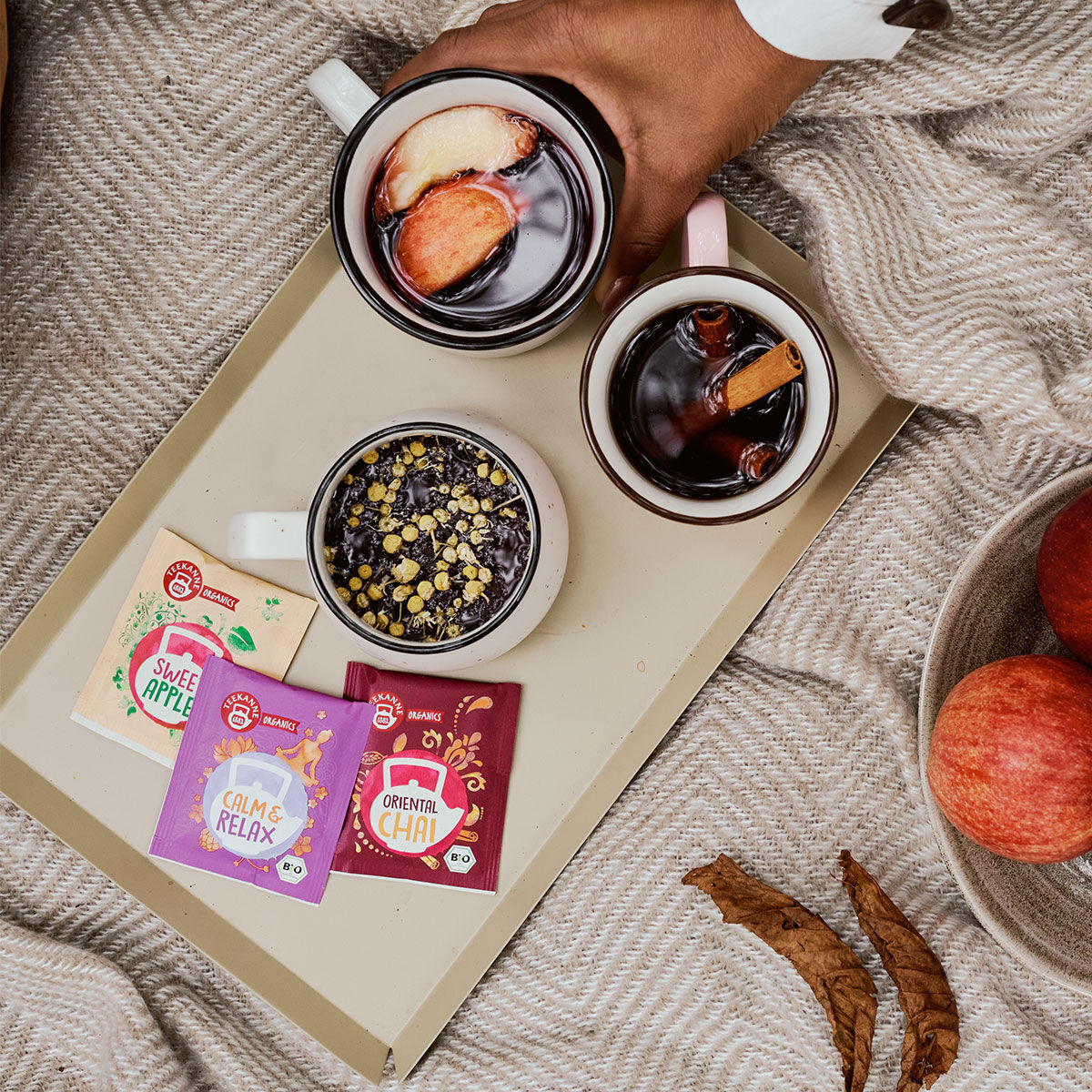Eine Hand hält eine Tasse Glühgetränk mit Fruchtscheiben neben zwei anderen Tassen auf einem Tablett mit ORGANICS Oriental Chai-Teepäckchen, einer Schale mit Äpfeln und einer kuscheligen Decke im Hintergrund.