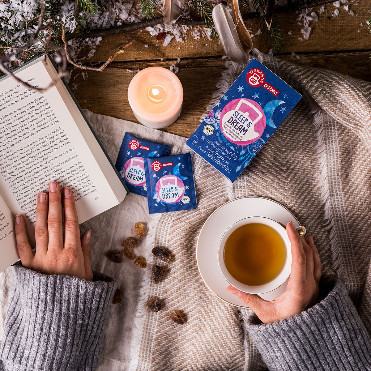 Eine Person in einem grauen Pullover liest ein Buch und hält dabei eine Tasse Bio-Kräutertee in der Hand. Daneben liegen ORGANICS Sleep & Dream Teebeutel, eine Kerze, Kandiszucker und Weihnachtsdekorationen auf einer gemütlichen karierten Decke.