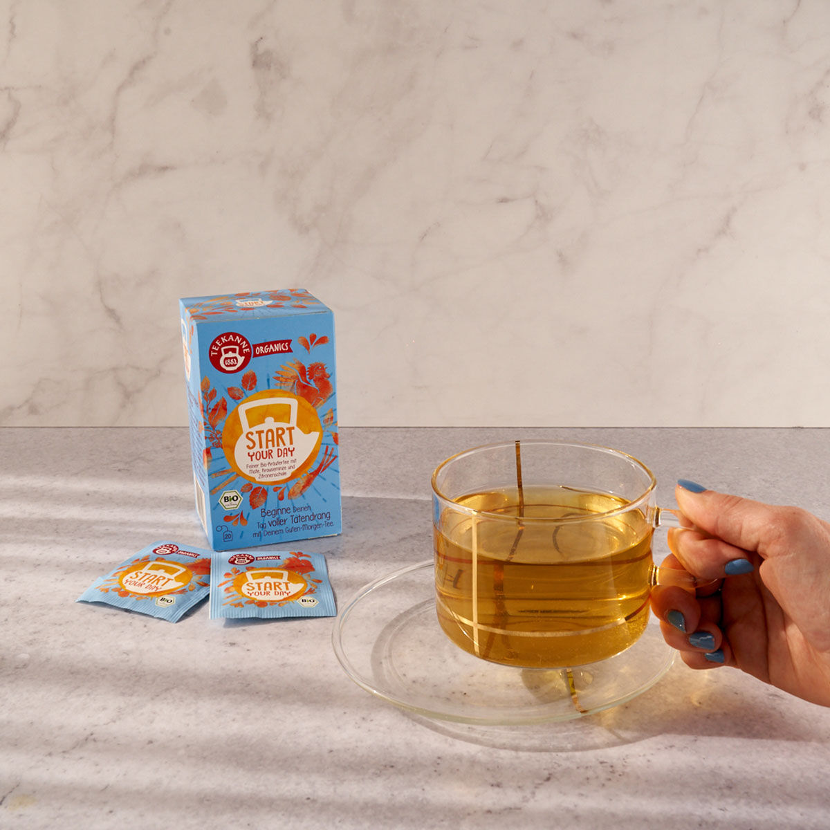 Eine Hand hält ein Glas mit hellem Tee auf einer Untertasse. Daneben stehen eine blaue ORGANICS Start Your Day-Box und zwei passende Teebeutel, alles arrangiert auf einer hellen Arbeitsplatte mit Marmorhintergrund.