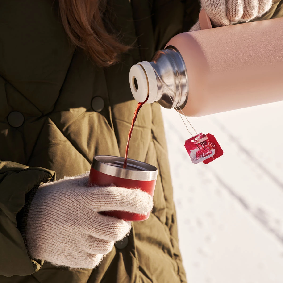 In einer grünen Steppjacke und beigen Handschuhen gießt jemand heißen Tee aus einer rosa Thermoskanne in eine rote Tasse. Auf den Teebeutelanhängern sind Schneeträumchen zu sehen, im Hintergrund liegt Schnee.