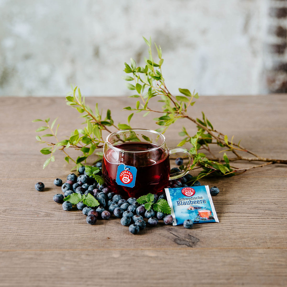 Auf einem Holztisch steht eine Tasse schwedischen Blaubeerentee, umgeben von frischen Blaubeeren, Minzblättern, einer Teebeutelpackung und grünen Zweigen im Hintergrund.