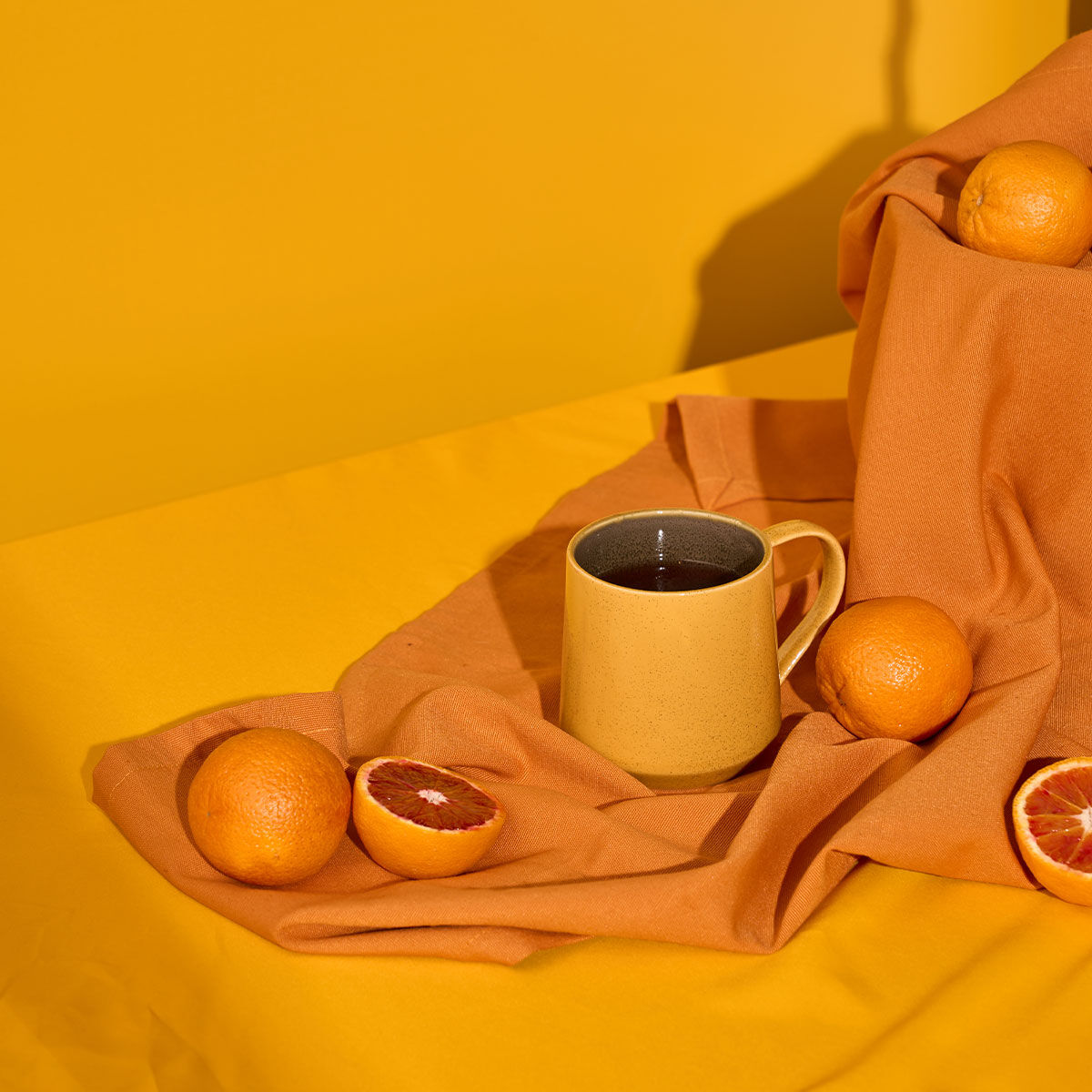 Eine beige Tasse mit dunklem Blutorangentee steht auf einem orangefarbenen Tuch, umgeben von ganzen und halbierten mediterranen Blutorangen vor einem leuchtend gelben Hintergrund.