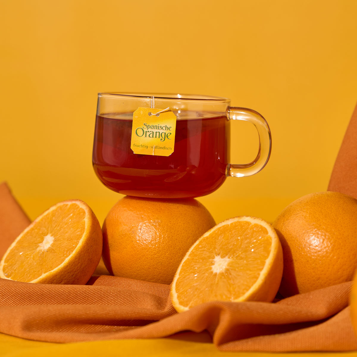 Eine Glastasse mit Spanischem Orangentee steht auf einer ganzen Orange, umgeben von einer halbierten und zwei ganzen Orangen, auf einem braunen Tuch mit orangefarbenem Hintergrund.